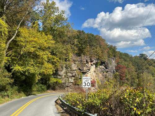 A winding road with a 30 mph speed limit sign, surrounded by colorful autumn trees and rocky cliffs under a blue sky.