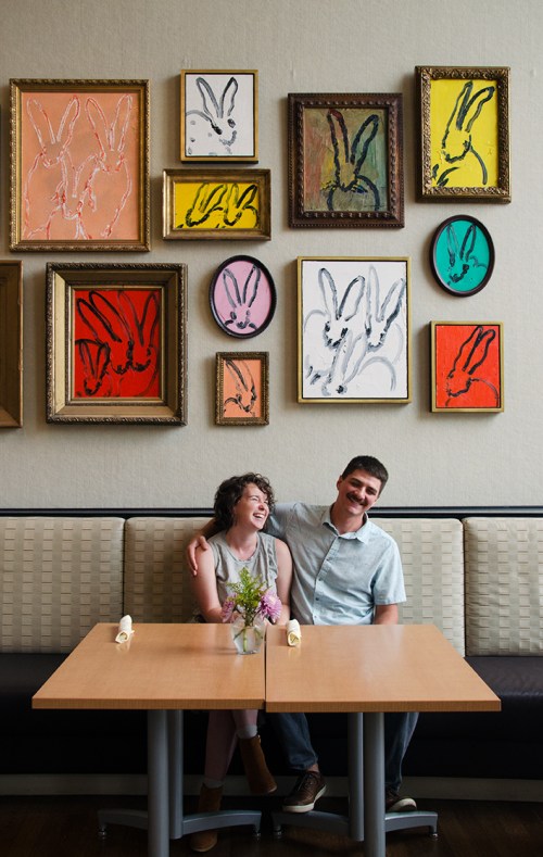 A couple sits at a table in a restaurant, smiling, with colorful bunny artwork displayed on the wall behind them.
