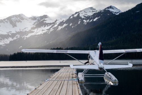 A seaplane is docked on a wooden pier, surrounded by mountains and a calm lake under a cloudy sky.