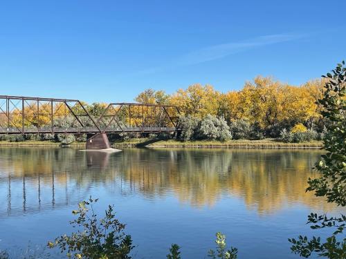 A tranquil river scene featuring a metal bridge and vibrant autumn trees reflecting in the water under a clear blue sky.