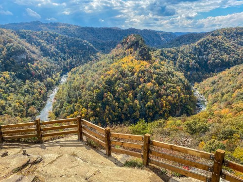 A scenic view of a river winding through colorful autumn hills, framed by a wooden railing under a cloudy sky.
