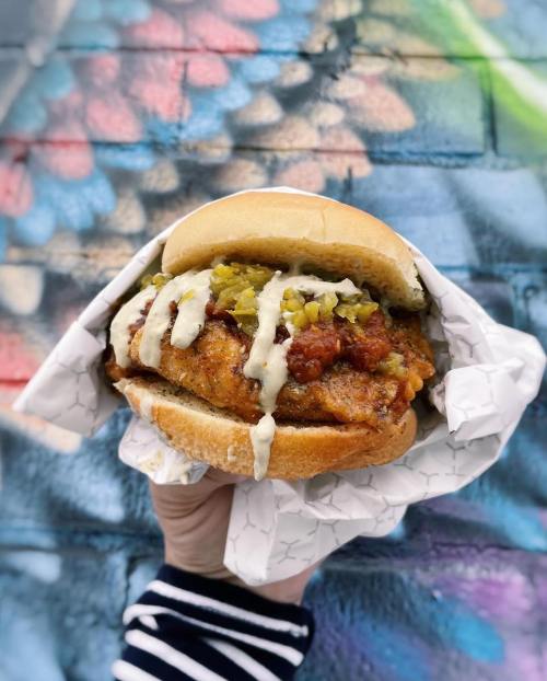 A hand holding a fried chicken sandwich with pickles and sauce, set against a colorful graffiti background.