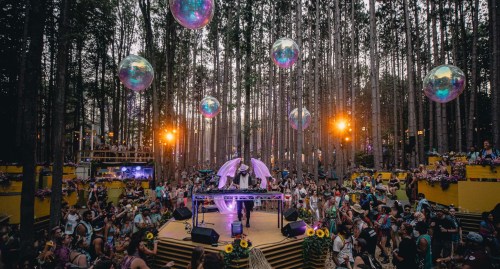 A vibrant outdoor music festival in a forest, featuring a DJ on stage with colorful lights and large bubbles overhead.