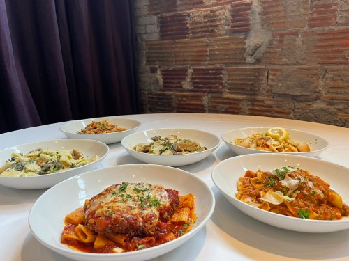 A table with six bowls of various pasta dishes, featuring different sauces and garnishes, against a rustic brick background.