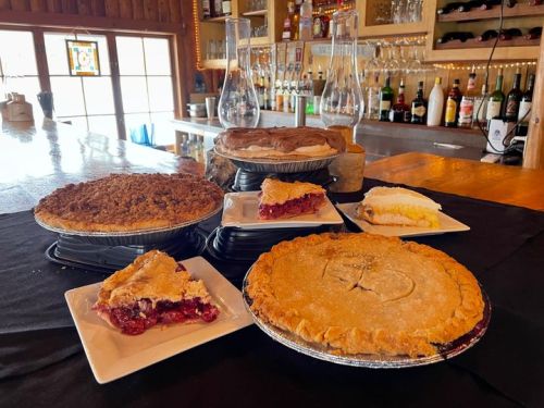 A variety of delicious pies displayed on a table, including cherry, apple, and cream pies, with a bar in the background.