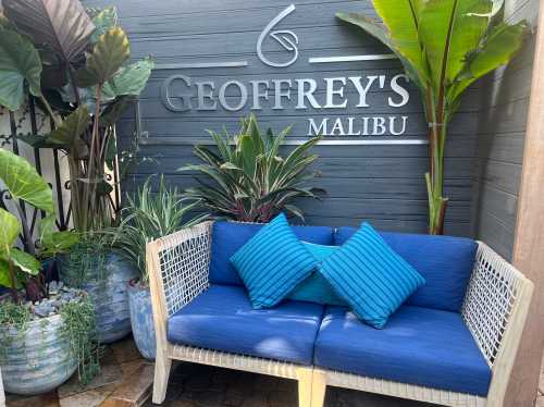 A cozy blue sofa with cushions in a tropical setting, surrounded by lush plants and a sign for Geoffrey's Malibu.