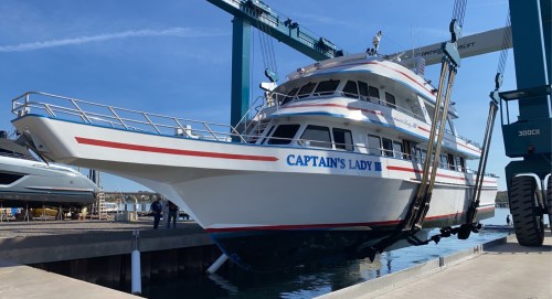 A large white and blue boat named "Captain's Lady III" being lifted by a crane at a marina.