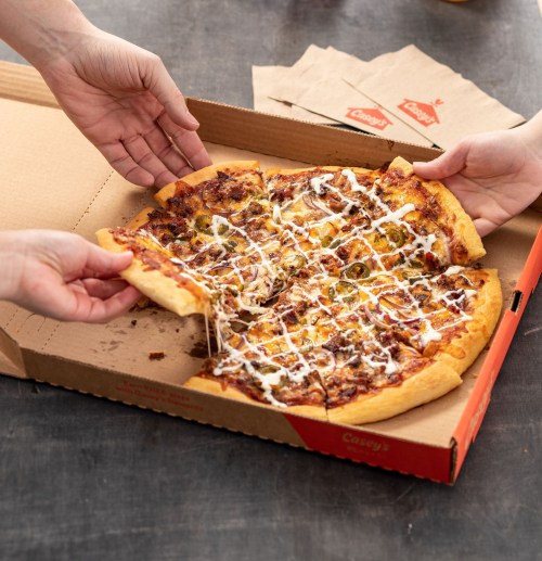 Two hands reaching for a slice of pizza with toppings and drizzled sauce, in an open pizza box.