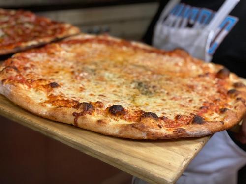 A person holds a large, freshly baked cheese pizza on a wooden board, with a golden crust and melted cheese.