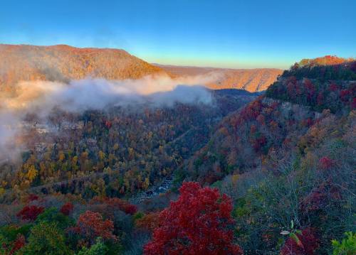 A scenic view of a valley with colorful autumn foliage and mist rising from the trees under a clear blue sky.