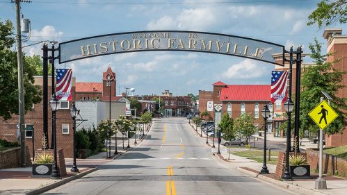 Welcome sign arching over a street in Historic Farmville, with buildings and flags lining the road under a blue sky.