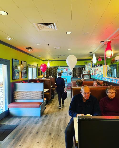 A colorful diner interior with booths, balloons, and patrons enjoying their meals. A person walks towards the exit.