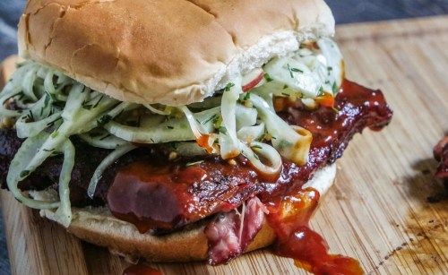 A close-up of a BBQ sandwich with pulled meat, topped with coleslaw, served on a wooden cutting board.