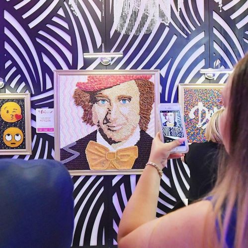 A person takes a photo of a colorful mosaic portrait of a man in a red hat and bow tie, displayed on a patterned wall.