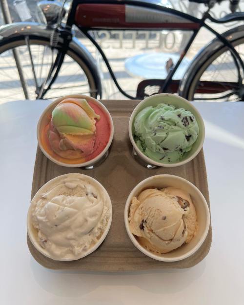 Four ice cream cups in a tray: pink, green, brown, and white flavors, with a bicycle in the background.