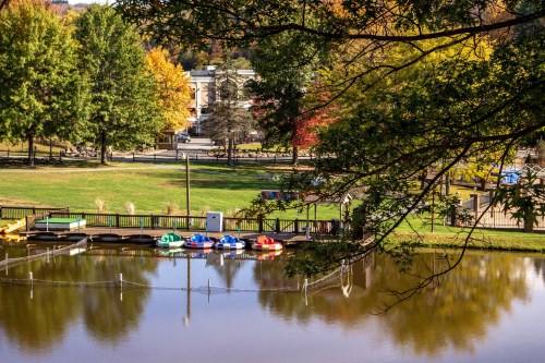 A serene park scene with colorful paddle boats on a calm pond, surrounded by trees and autumn foliage.