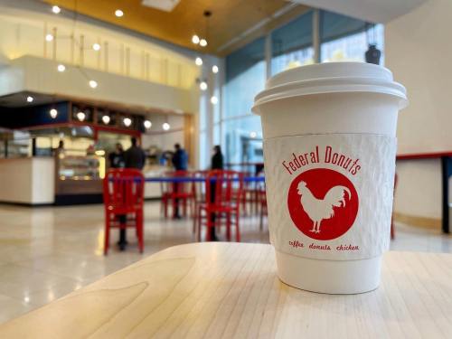 A coffee cup from Federal Donuts sits on a table in a bright, modern café with red chairs and a donut display in the background.