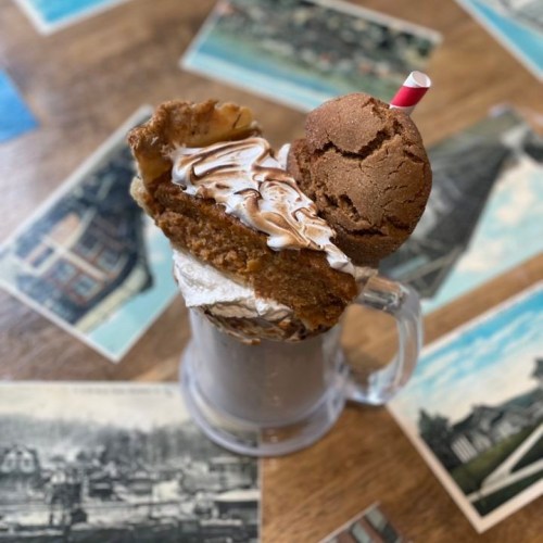 A tall glass filled with chocolate milkshake, topped with whipped cream, cookie, and a slice of cake, on a vintage table.