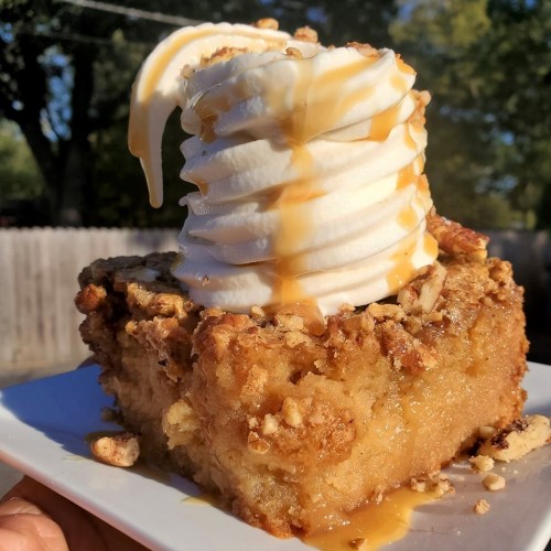 A slice of dessert topped with whipped cream and caramel drizzle, served on a white plate outdoors.
