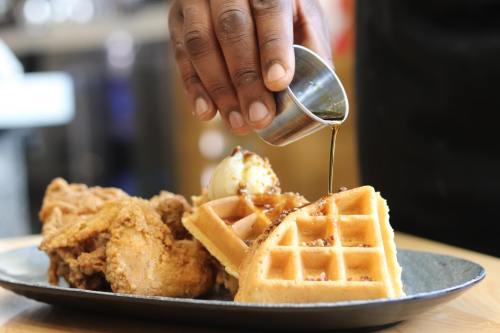 A hand pours syrup over a waffle, accompanied by fried chicken and a scoop of butter on a black plate.