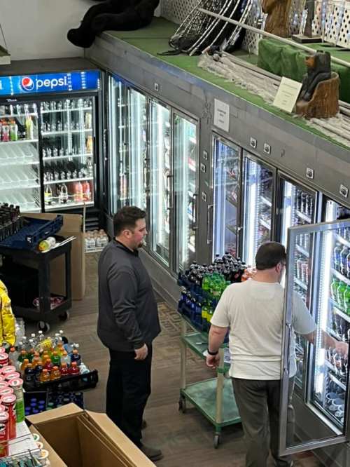 Two men browse a beverage aisle in a store, surrounded by coolers filled with various drinks.