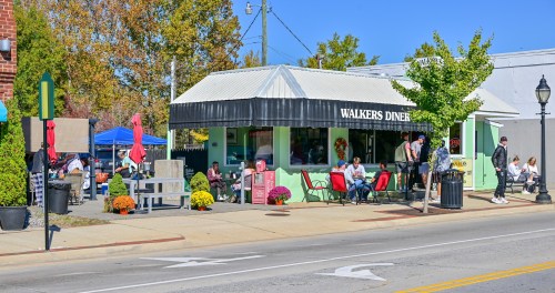A colorful diner with outdoor seating, surrounded by trees and flowers, on a sunny day. People are dining outside.