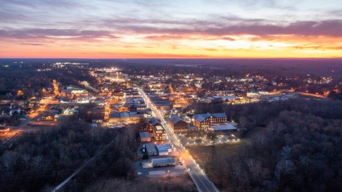 Aerial view of a small town at sunset, with streets illuminated and a vibrant sky in the background.