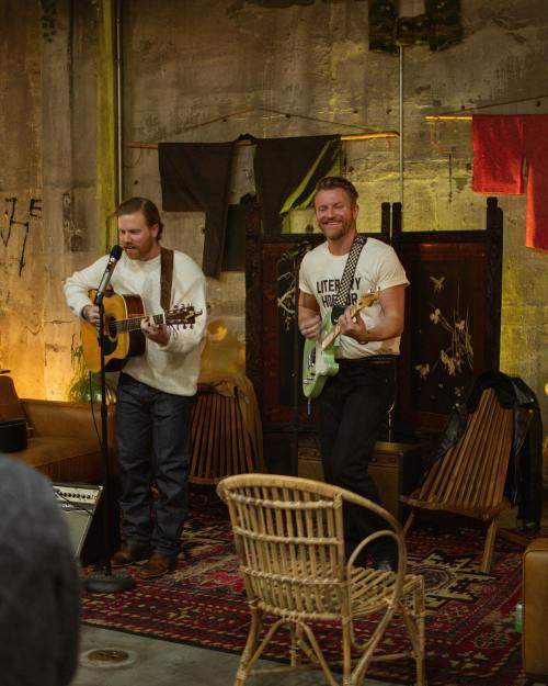 Two musicians perform in a cozy, rustic space, one with an acoustic guitar and the other with an electric guitar.