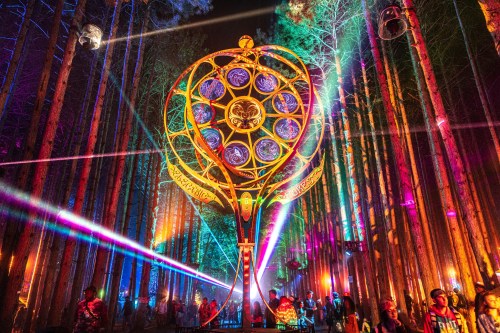 A vibrant forest scene illuminated by colorful lights and lasers, featuring a large decorative structure among trees.
