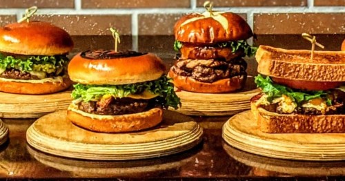 A variety of gourmet burgers on wooden plates, featuring different toppings and styles against a brick background.
