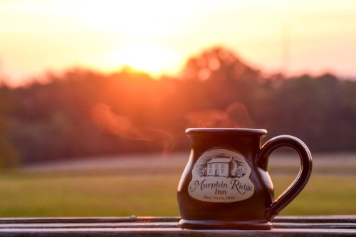 A steaming mug sits on a wooden surface with a sunset in the background, casting warm colors over the landscape.