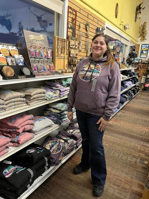 A woman stands in a store aisle, smiling beside shelves of clothing and souvenirs, with various items displayed around her.
