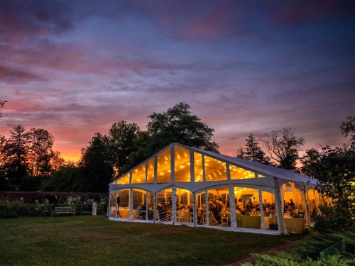 A beautifully lit tent at sunset, filled with guests, surrounded by trees and a colorful sky.