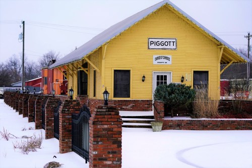 A yellow train station building with "Piggott" sign, surrounded by snow and brick fencing, on a cloudy day.