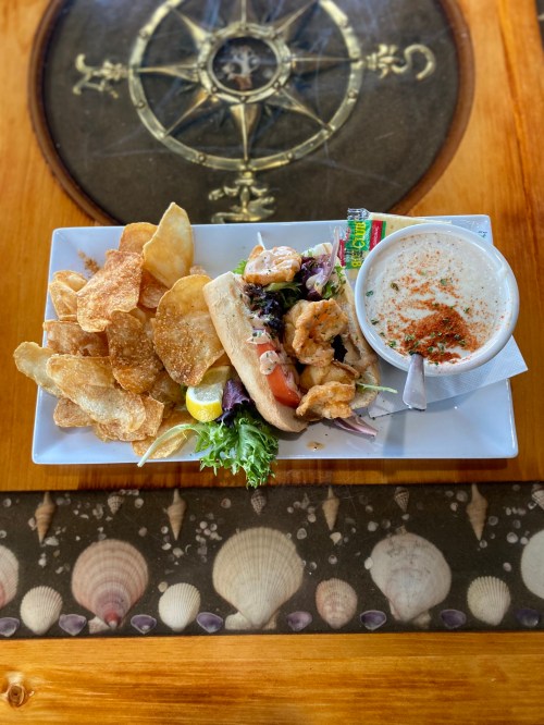 A plate featuring a shrimp sandwich with lettuce and tomato, a bowl of soup, and a side of crispy potato chips.