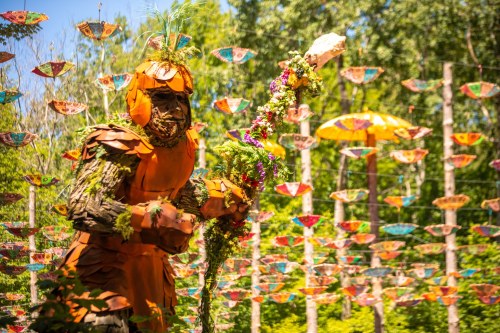 A large, whimsical figure made of natural materials stands amidst colorful hanging decorations in a forest setting.