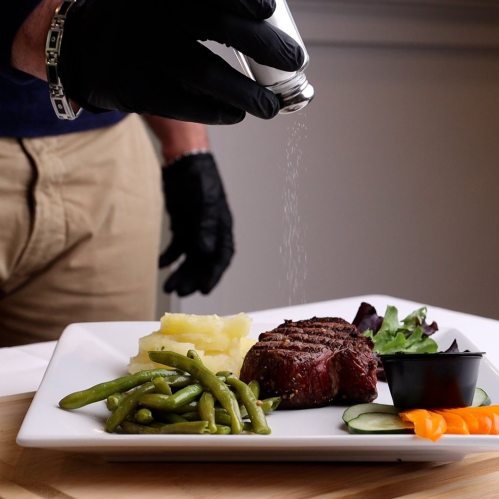 A person in black gloves seasons a plated steak with salt, accompanied by green beans, mashed potatoes, and salad.