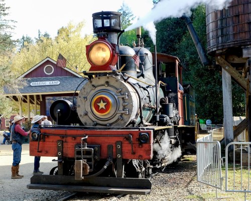 A vintage steam locomotive with a red and black design, surrounded by trees and a boarding camp building in the background.