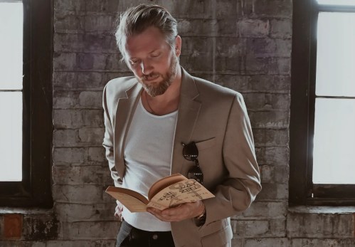A man in a blazer reads a book while standing in a room with exposed brick walls and large windows.