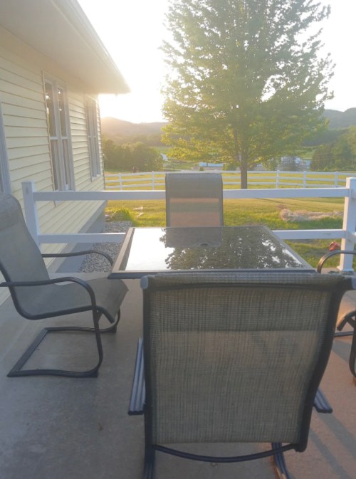 A patio with a glass table and two chairs, overlooking a green landscape and a tree at sunset.