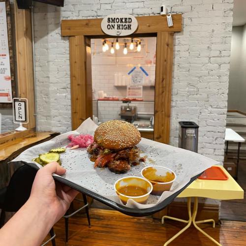 A hand holds a tray with a sesame seed burger, pickles, and two dipping sauces in a casual dining setting.