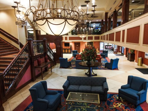 A spacious hotel lobby with blue chairs, a large chandelier, and decorative plants, featuring a staircase in the background.