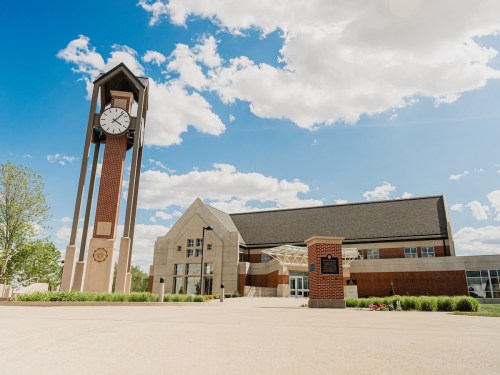 A tall clock tower stands beside a modern building under a blue sky with fluffy clouds. Greenery is visible in the foreground.