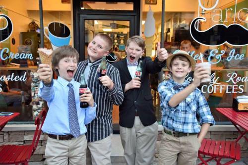 Four boys in dress shirts and slacks pose excitedly outside an ice cream shop, holding drinks and ice cream cones.