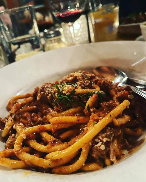 A close-up of a plate of pasta with a rich sauce, garnished with herbs, and glasses of drinks in the background.