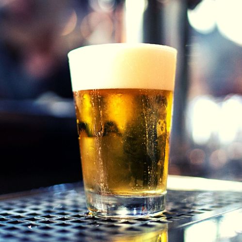 A cold glass of beer with a frothy head, sitting on a bar counter with a blurred background.