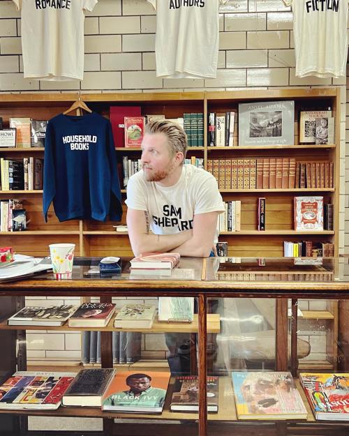 A man in a t-shirt leans on a counter in a cozy bookstore filled with books and shelves.