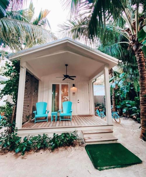 A charming beach cottage with turquoise chairs on a porch, surrounded by lush palm trees and sandy pathways.