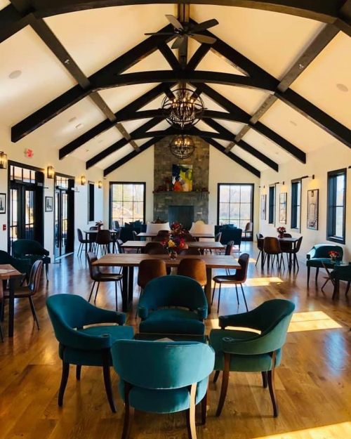 A spacious, modern room with wooden beams, a stone fireplace, and stylish chairs arranged around tables.