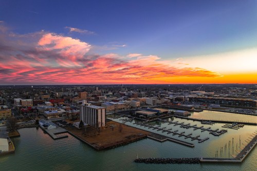 Aerial view of a city at sunset, featuring a marina, buildings, and colorful clouds in the sky.